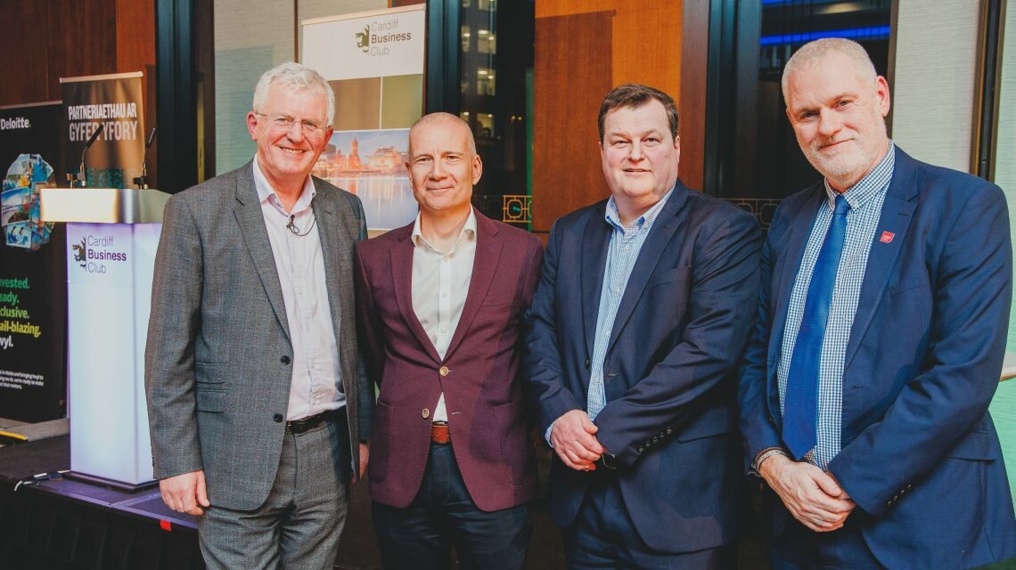 Scott Waddington, Ian Howse, Gareth Williams and Dr Ben Calvert, all dressed in suits, stand in front of a stage. In the background is a Cardiff Business Club branded lectern and banners.