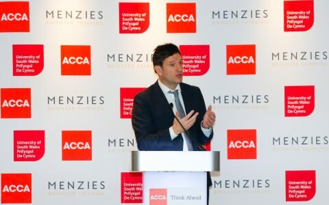 Noel Mooney stands behind a lectern and speaks to guests at ACCA's St David's Day Breakfast. He is standing in front of a backdrop with ACCA, Menzies and University of South Wales logos on it